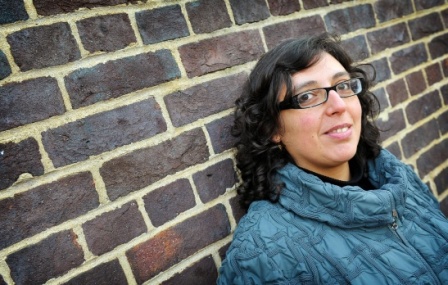 Artist Ana Laura Lopez wearing a blue jumper and leaning on a brick wall