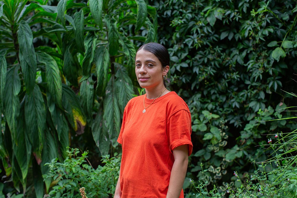 Artist Meera Shakti Osbourne standing in front of trees wearing an orange t-shirt