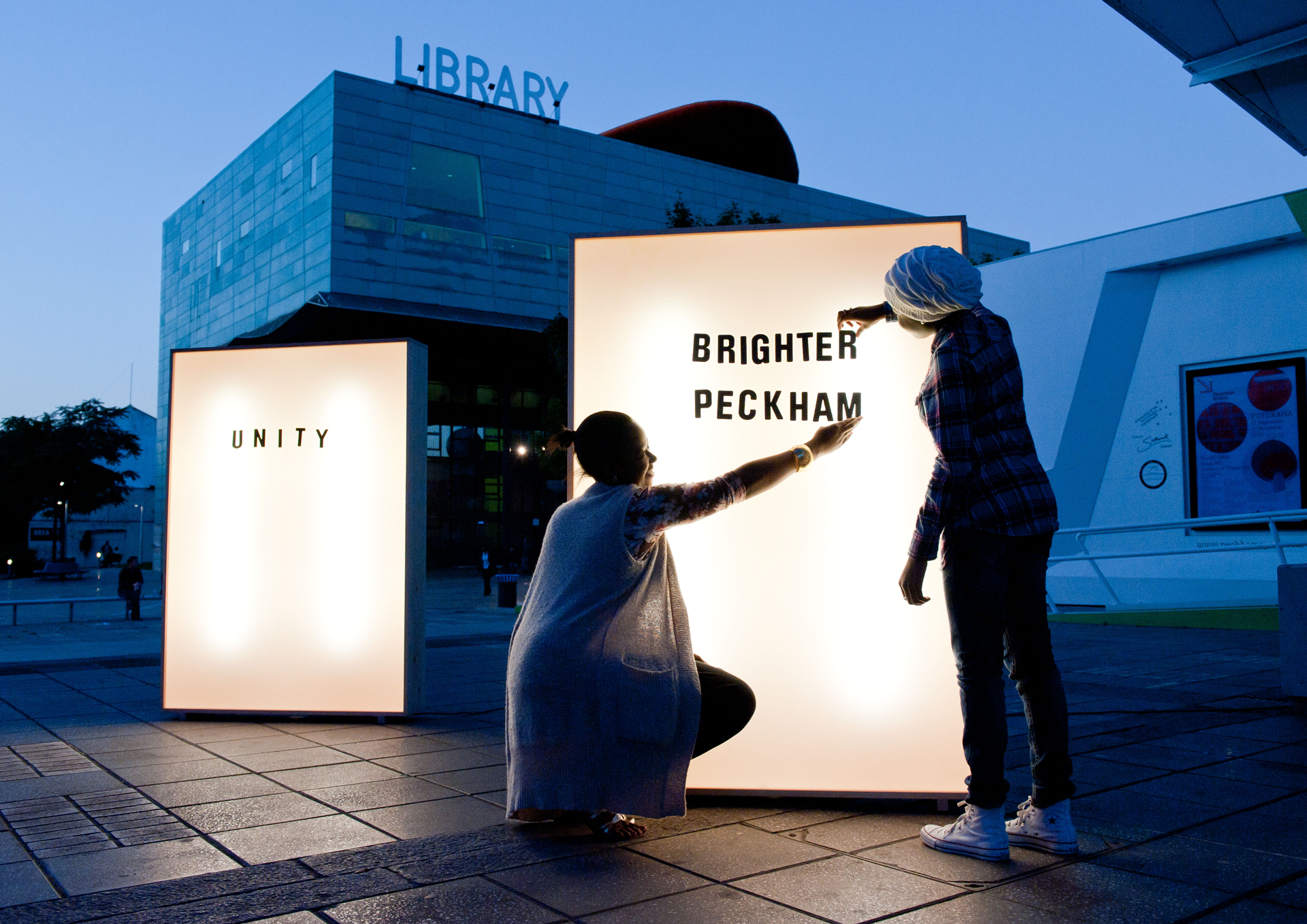 Two people adding black letters to a lighbox that reads Brighter Peckham outside Peckham library