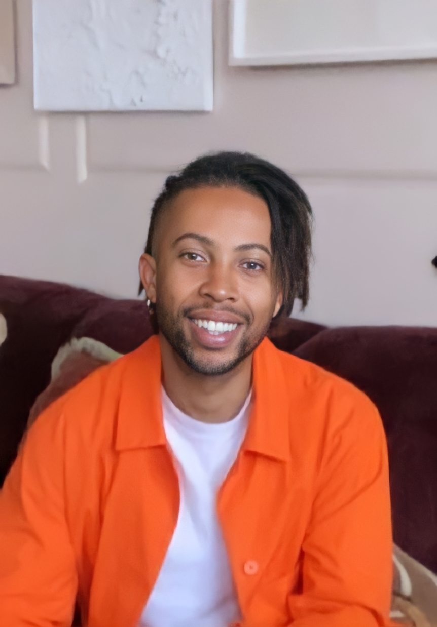 Brian Maina. A smiling young man with a side part and undercut dreds wearing a vibrant orange shirt over a plain white tee.