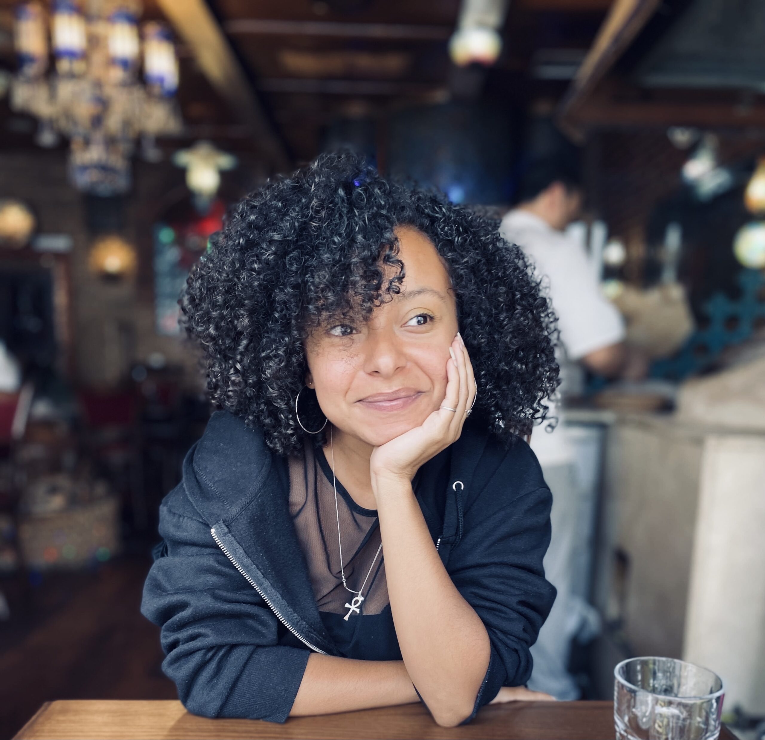 A portrait of a young woman with mixed heritage and large dark 3A curls cupping her face with one hand and smiling at something off camera.