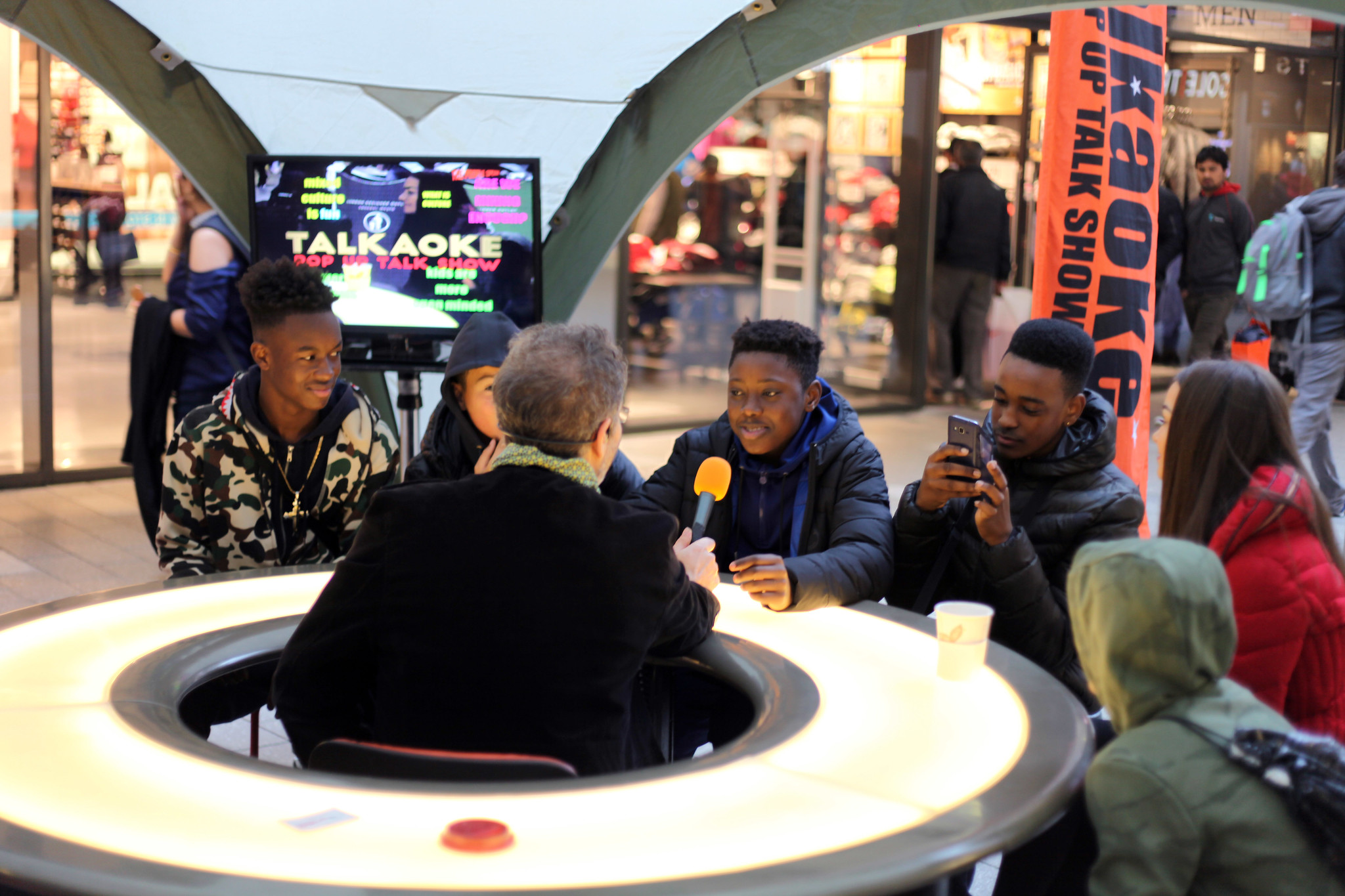 A group of young people sit at a roundtable with a host in the middle holding a microphone on his hand