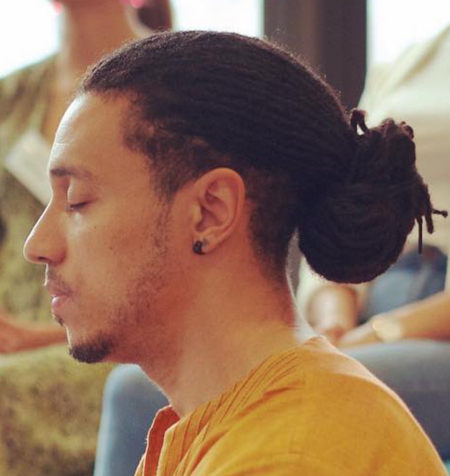 A portrait of a young man in profile with his eyes peacefully closed and his locs held back in a bun.